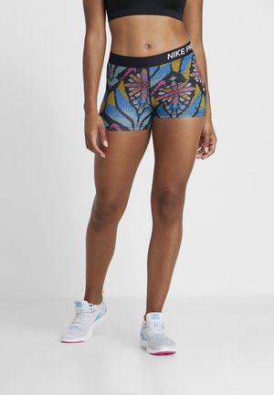 Femme portant un brassard de sport noir, un short Nike Pro à motif floral multicolore, et des chaussures de sport blanches avec des accents bleus et roses.
