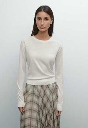 Pull long beige clair à manches longues avec un col rond et un ourlet côtelé, associé à une jupe à carreaux dans des tons doux de beige et de marron.