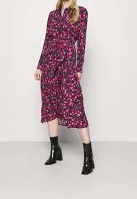 Frau in einem langärmeligen, pinken und lila Blumenkleid mit Midilänge, einem Taillengürtel und schwarzen, hochhackigen Stiefeletten, die vor einem schlichten Hintergrund steht.