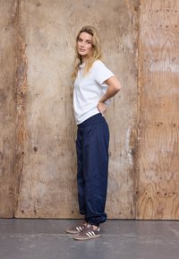 Jauna moteris su ilgais blondinais plaukais, vilkinti baltą marškinėlį, plačias tamsiai mėlynas kelnes ir rudus sportbačius, stovinti rankomis ant klubų priešais medinę sieną.