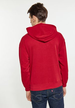 Rode hoodie met een relaxte pasvorm, geribde manchetten en tailleband. De stof lijkt zacht met een gladde textuur. Gepaard met donkere spijkerbroeken.