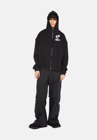 Haut à zip noir avec logo, associé à un pantalon cargo noir muni de plusieurs poches et de bas ajustables. Fini mat, coupe décontractée.