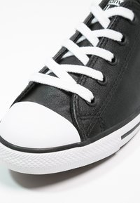 Baskets basses noires avec embout en caoutchouc blanc, lacets blancs et semelle blanche texturée sur fond blanc.