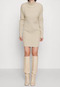 Strickkleid in hellbeige mit hohem Ausschnitt und geripptem Muster, kombiniert mit kniehohen beigen Stiefeln, die beide ein schlankes Design aufweisen.
