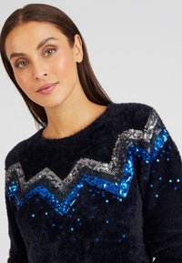 Navyblauer, kuscheliger Pullover mit einem Zickzackmuster aus silbernen und blauen Pailletten auf der Brust, mit einer strukturierten und weichen Oberfläche.