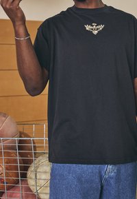 Czarna bawełniana koszulka z okrągłym dekoltem, zdobiona złotym emblematem na klatce piersiowej. Połączona z niebieskimi dżinsowymi szortami i otoczona piłkami sportowymi.