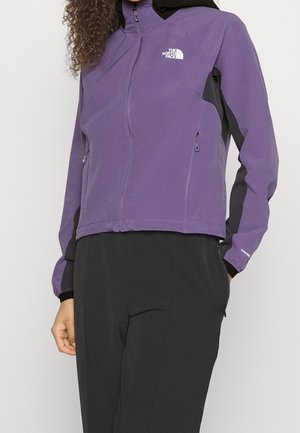 Personne portant une veste zippée The North Face violette avec des panneaux noirs et un pantalon noir, debout les mains détendues le long du corps.