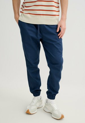 Hombre con pantalones deportivos azul marino, zapatillas blancas con suelas beige y un suéter crema con rayas naranjas, de pie sobre un fondo blanco.