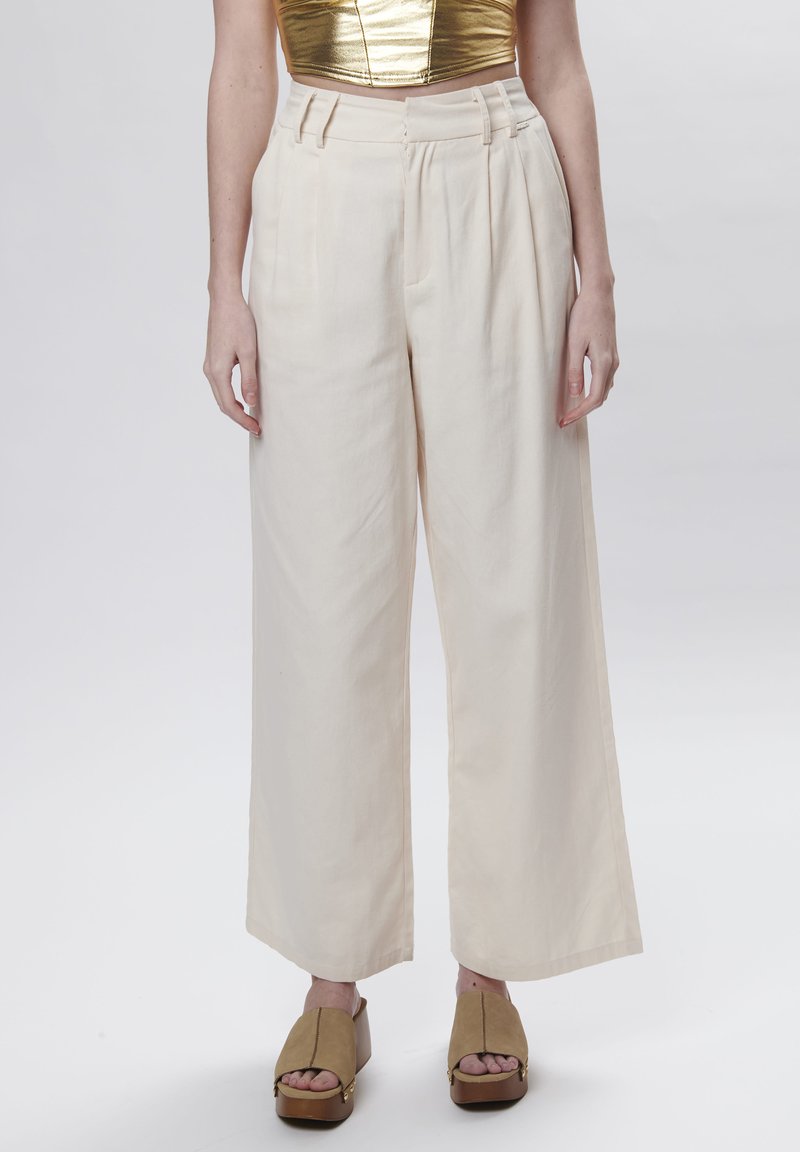 Pantalones de pierna ancha en beige con pliegues, hechos de una tela ligera. Presentan una cintura alta y trabillas para cinturón. Combinados con sandalias de plataforma marrón.