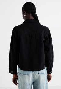 Mustattu farkkutakki napillisilla hihoilla ja klassisella kauluksella, päällä vaaleansiniset farkut, esittäen puhdasta ja minimalistista muotoilua.