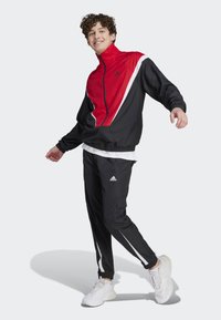 Tuta adidas nera e rossa con accenti bianchi, giacca con zip, vita elastica, pantaloni slim-fit con tasche cargo e sneakers bianche.