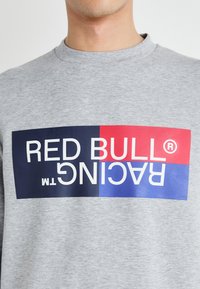 Siva dukserica s velikim pravokutnim grafičkim dizajnom na kojem piše "RED BULL® RACING" u bijelom tekstu, istaknuta crvenim i plavim dijelovima.