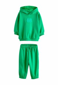 Zöld pulóver kapucnis felső elülső grafikus szöveggel, hozzá illő zöld melegítőnadrággal, amely elasztikus derékpánttal és bordázott mandzsettákkal rendelkezik.