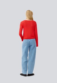 Red ribbed sweater with long sleeves worn over light blue loose-fitting jeans. Black shoes visible; model's back facing the camera.