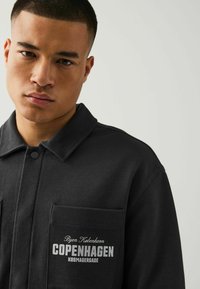 Schwarz strukturierte Jacke mit einem geknöpften Kragen, einer Brusttasche mit dem grafischen Text "KOPENHAGEN" und einer glatten Stoffoberfläche.