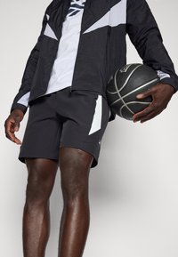 Basketbalový outfit s černou bundou s bílými akcenty, bílým tričkem pod ní a černými šortkami. Model drží basketbal.