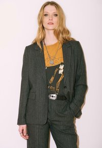 Femme aux longs cheveux blonds portant un costume gris à rayures, un t-shirt graphique de groupe moutarde, un collier en argent et une ceinture noire avec une boucle argentée.