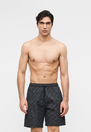 Homme torse nu aux cheveux courts et foncés, portant un short de bain noir à motifs avec cordon de serrage, debout devant un fond clair uni.