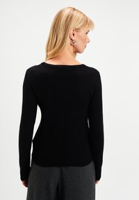 Blondynka stojąca bokiem, nosząca srebrne kolczyki w kształcie kół, czarną bluzkę z długim rękawem i szare spodnie na tle białego tła.