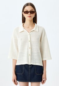 Kortærmet, knappet skjorte i hvidt hæklet strik. Har en krave-udskæring og en afslappet pasform, parret med en denim mini nederdel.