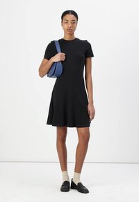 Musta lyhythihainen mekko, jossa on istuva bodice ja levenevä hame. Vaatetta on täydentämässä sininen olkalaukku, beige sukat ja mustat liukuhihnat.