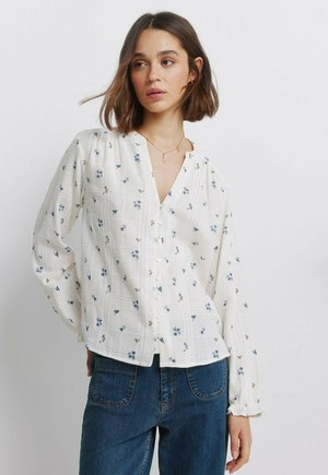 Nuori nainen, jolla on olkapäille ulottuvat ruskeat hiukset ja joka on pukeutunut valkoiseen nappipaitaan, jossa on pieniä sinisiä kukkakuvioita, sekä sinisiin farkkuihin.