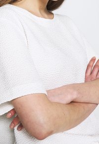 Vit texturerad topp med rund halsringning och korta ärmar, med ett vävt tygmönster. Armarna korsade i en neutral pose.