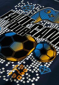 Camiseta azul marino con un diseño gráfico que presenta pelotas de fútbol en negro, azul y amarillo, patrones en píxeles y un texto en negrita que dice "FAULT GAME YOUR."