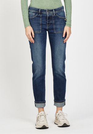 Henkilö, jolla on ylhäältä rullatut tummansiniset farkut, vaaleanvihreä pitkähihaisen paita ja valkoiset lenkkarit, seisomassa yksiväristä taustaa vasten.