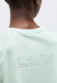 Εγγύς λήψη του λαιμού και του άνω μέρους της πλάτης ενός ατόμου που φοράει ένα ανοιχτό πράσινο πουκάμισο από διάτρητο ύφασμα με την επιγραφή "LDN" σε ανακλαστικά γράμματα.