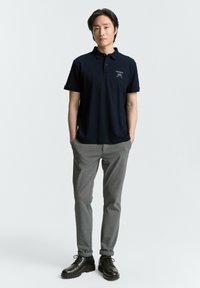 Marineblauwe polo met korte mouwen, voorzien van een geborduurd logo op de borst, gecombineerd met lichtgrijze taps toelopende broeken en zwart veter schoenen.