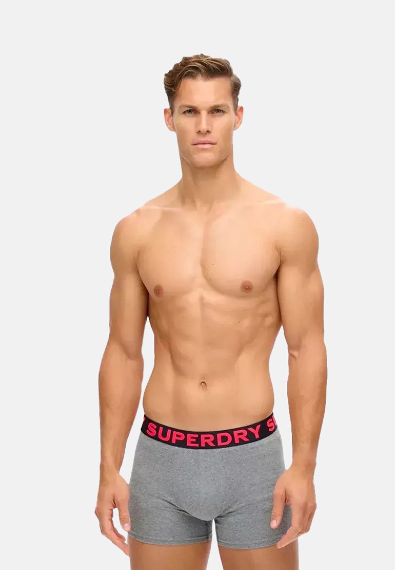 Bóxer gris con una cinturilla negra que presenta "SUPERDRY" en letras grandes de color rosa. El material parece suave con un diseño ajustado.