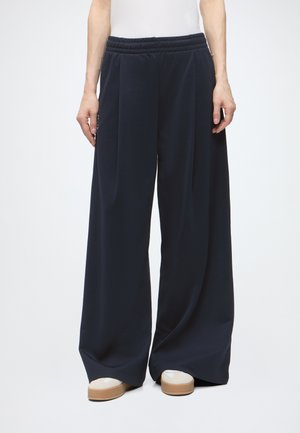 Persoană purtând pantaloni largi bleumarin și adidași albi cu tălpi bej, stând în fața unui fundal alb simplu.