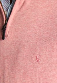 Detailný záber na ružový sveter so štvrťzipsom a malým vyšívaným logom zajaca na hrudníku, pod ktorým je pruhovaná košeľa.