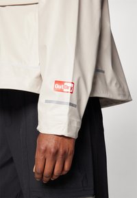 Manche de veste imperméable beige avec logo "OutDry" visible en rouge. Présente des accents réfléchissants et un design de tissu texturé.