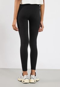 Colanți negri dintr-un material elastic, cu un design ajustat și talie înaltă, având o textură netedă. Purtați împreună cu încălțăminte sport albă și gri.