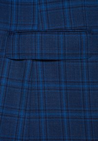 Niebieska tkanina w kratę z kieszenią z klapą. Wzór obejmuje ciemnonawigacyjne i jaśniejsze niebieskie linie krzyżujące się w układzie siatki.