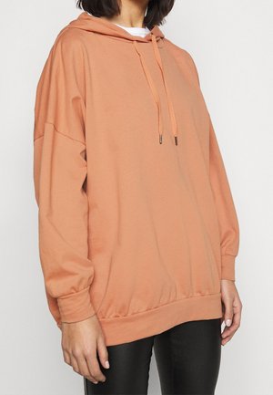 Osoba ubrana w oversize'ową brzoskwiniową bluzę z kapturem z troczkami i czarne spodnie, przedstawiona od szyi do połowy uda na jednolitym tle.