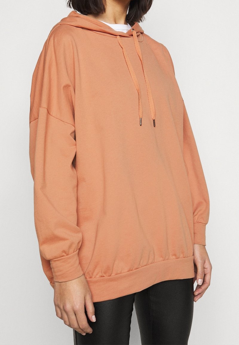 Personne portant un sweat à capuche pêche surdimensionné avec cordons de serrage et pantalon noir, visible du cou jusqu'au milieu des cuisses sur un fond uni.