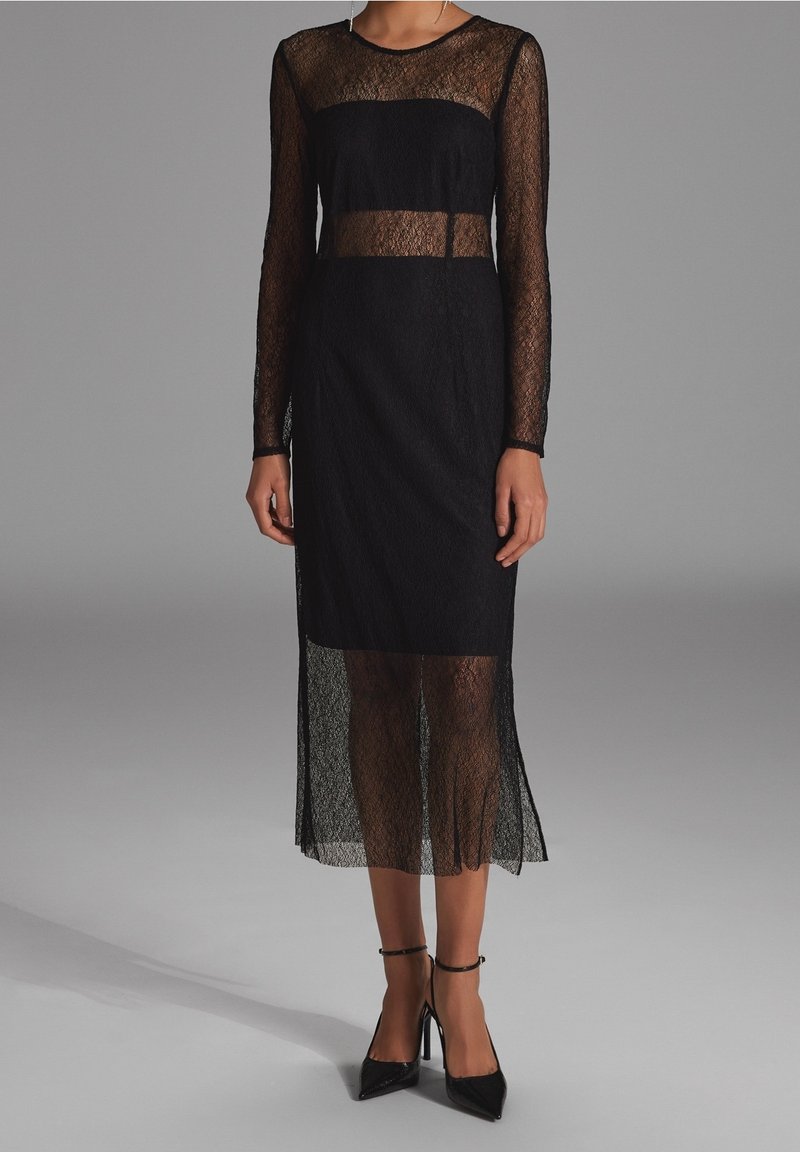 Femme portant une robe midi en dentelle noire avec des manches longues transparentes et des panneaux transparents à la taille et à l'ourlet, associée à des talons noirs à bride de cheville.