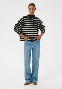 Zwart-witte gestreepte cardigan met een hoge nek, knopen aan de voorkant en een ontspannen pasvorm. Gecombineerd met lichtblauwe wijde jeans en bruine puntige ballerina's.