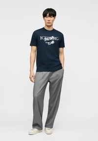Homme portant un T-shirt graphique bleu marine avec texte blanc, pantalon plissé gris et baskets blanches, debout avec une main dans la poche.