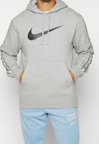 Grå sweatshirt i en bomullsblandning, med en svart Nike-logotyp, dragsko i huven och kontrasterande sidostreck i vitt. Framficka ingår.
