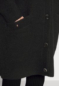Main portant une manche tricotée noire reposant dans la grande poche avant d'un cardigan en laine noir boutonné, porté sur un pantalon noir côtelé.