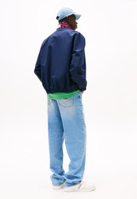 Dunkelblaue Jacke mit elastischem Bund, grünem Shirt darunter, hellblauen Jeans und weißen Sneaker. Enthält eine Baseballkappe.