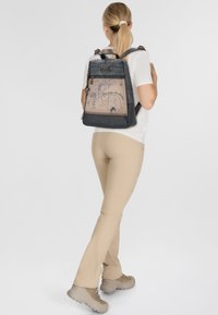 Mochila con material texturizado en azul oscuro, parte frontal en beige claro con diseños ilustrados y correas ajustables dobles. Compartimento con cremallera visible.