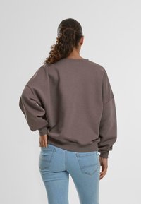 Felpa grigia oversize con spalle scese e polsini a coste, abbinata a jeans chiari a vita alta, vista da dietro.