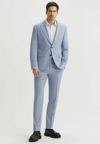 Jasnoniebieski garnitur z gładkiego materiału, z klapami typu notch, jednorzędowy, w połączeniu z czarnymi butami. Widoczna biała koszula w kratę.