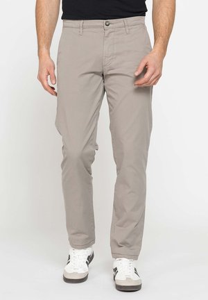 Uomo che indossa pantaloni beige a gamba dritta e scarpe da ginnastica bianche con strisce nere, in piedi su sfondo bianco.