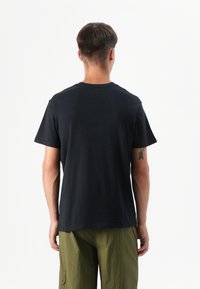 Personne portant un t-shirt noir et un pantalon vert olive, vue de dos sur un fond blanc uni.